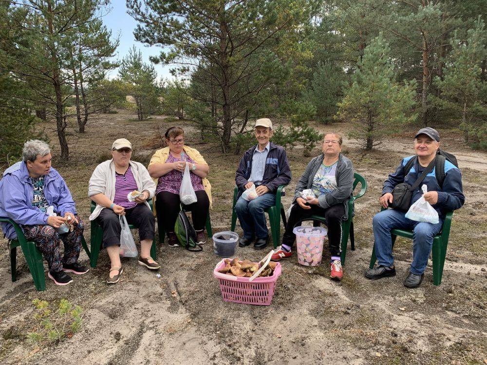 Grupa mieszkańców siedzi na krzesełkach w lesie i spożywają posiłek. 