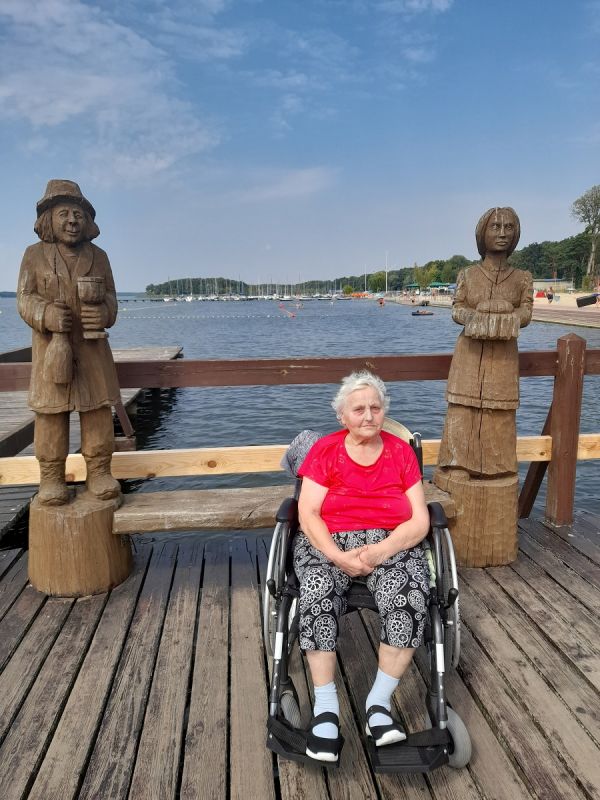 Mieszkanka na wózku inwalidzkim. W tle jezioro, a po bokach figury z drewna. 