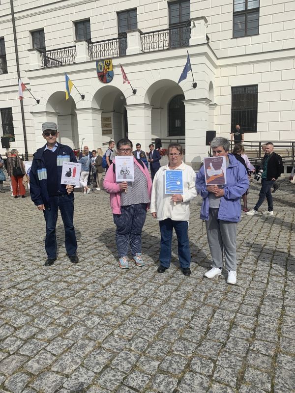 Miszkańcy stoją z transparentami o tematyce niepełnosprawności, w tle głogowski Ratusz. 