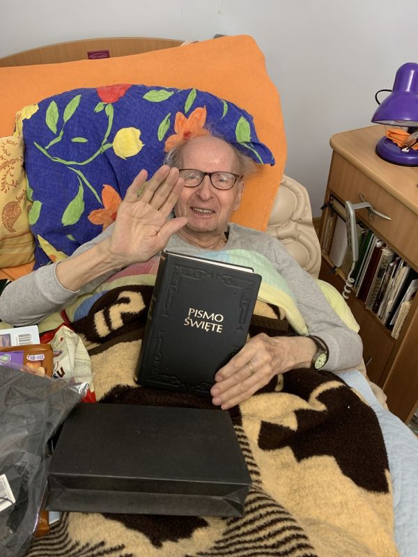 Mieszkaniec leżący w łóżku trzyma biblię w ręku i macha ręką 