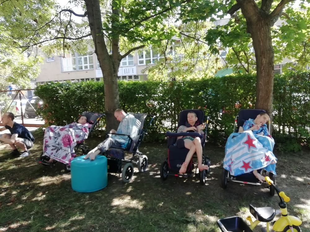 Podopieczni siedzą w wózkach inwalidzkim w ogrodzie